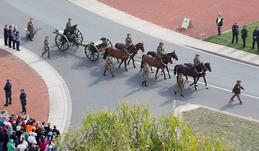 An 18-pounder field gun and limber drawn by a team of horses, escorted by soldiers in period uniform through a street lined with spectators.